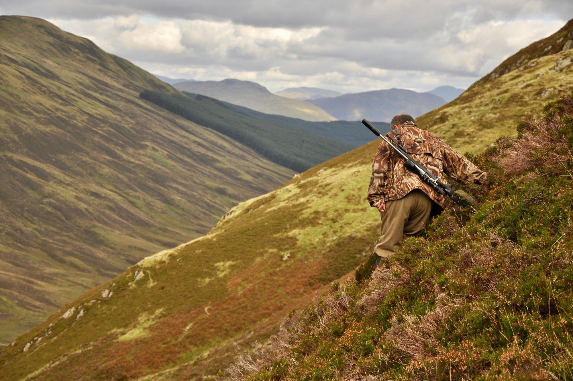 A hunter in camouflage climbing a steep grassy mountainside with a rifle, a long valley below.